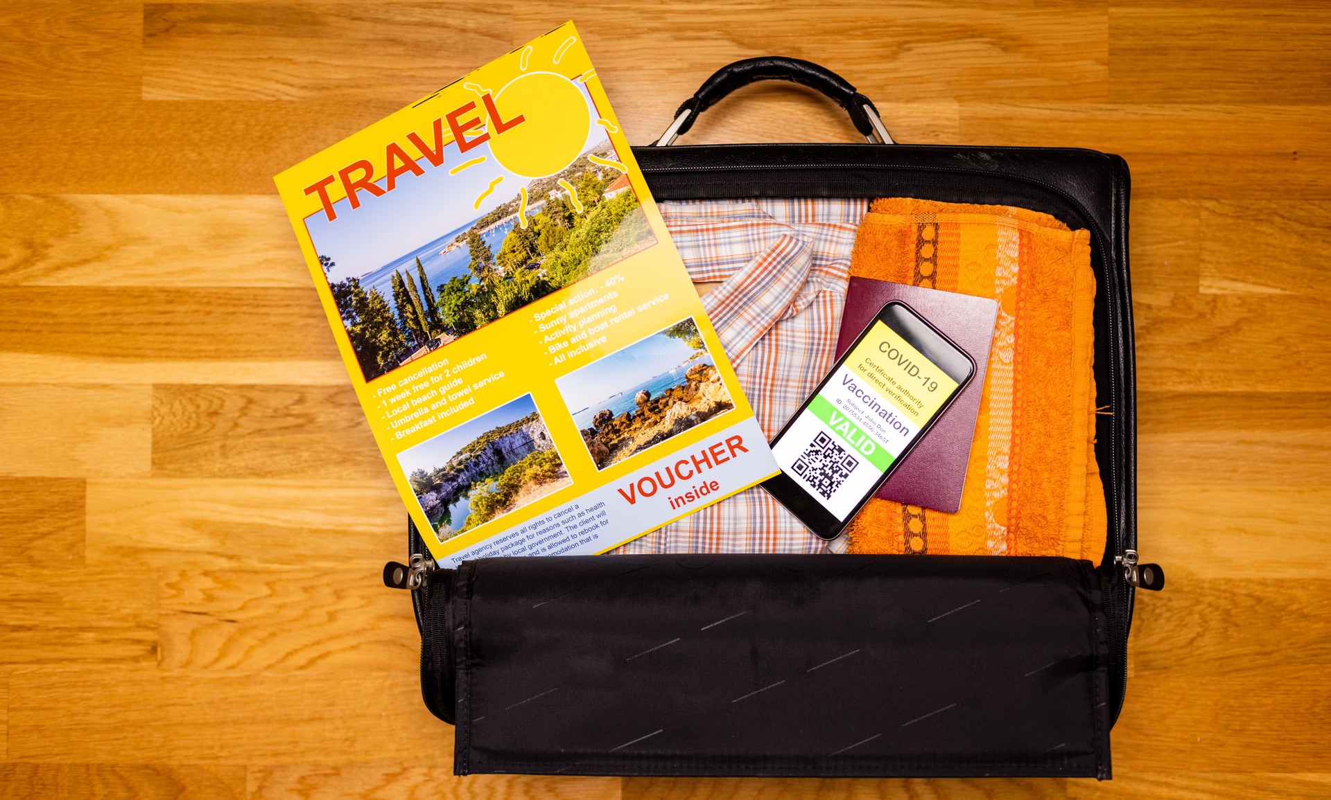 Suitcase with travel brochure and mobile phone showing vaccination certificate along with paper passport. Copyspace on wooden floor