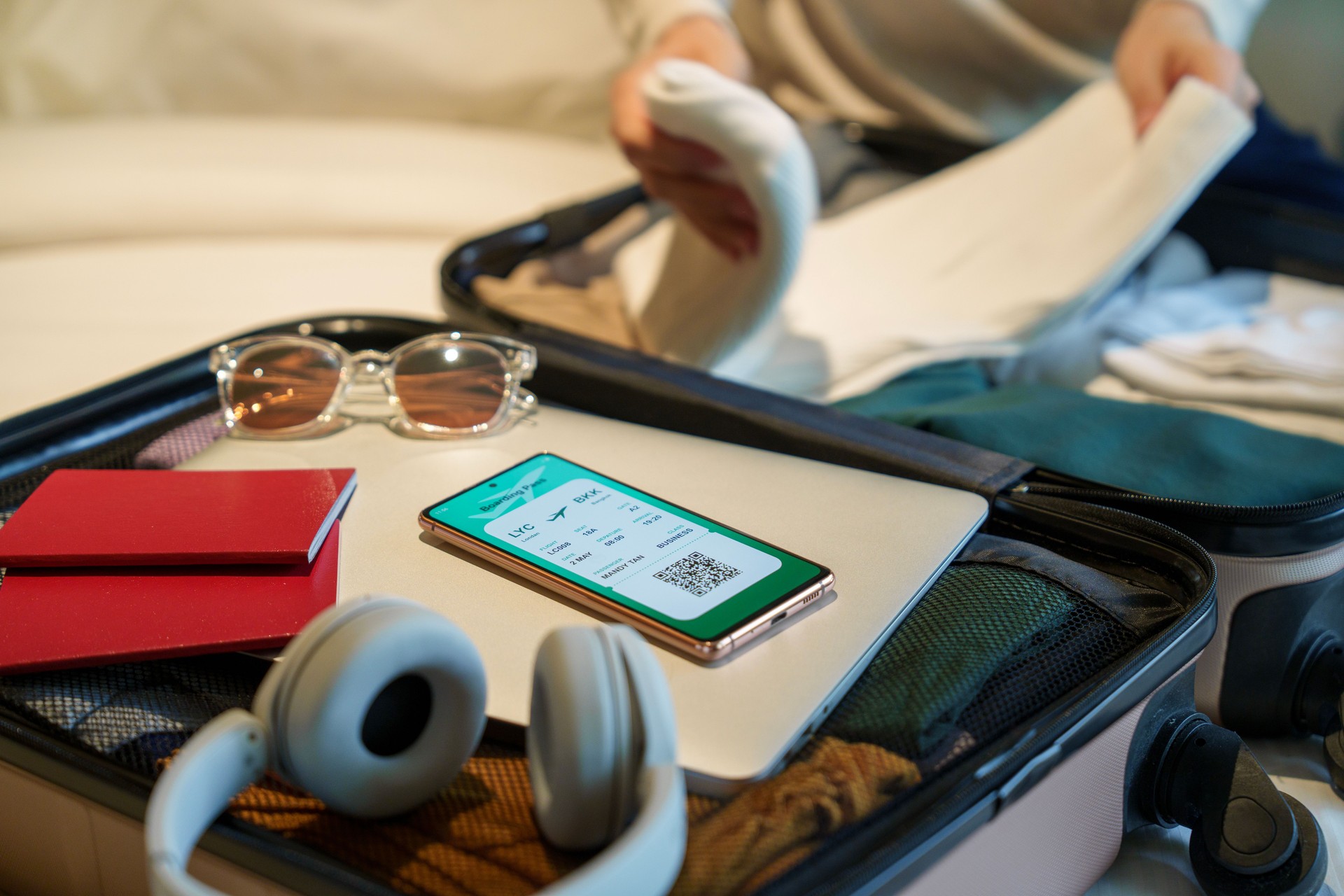 Close-up image of a smartphone displaying an electronic flight ticket next to a woman's packed suitcase containing clothing, sunglasses, headphones, a laptop, and a passport, placed on a bed.